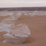 A breathtaking photo of white chalk rock formations rising like giant stone mushrooms from the pale sand in Sahara El Beyda under a bright blue sky.