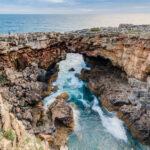 A dramatic wide-angle shot of Boca do Inferno in Cascais, Portugal, showing crashing Atlantic waves splashing against the natural limestone rock arch under a cloudy sky.