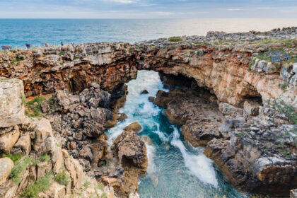 A dramatic wide-angle shot of Boca do Inferno in Cascais, Portugal, showing crashing Atlantic waves splashing against the natural limestone rock arch under a cloudy sky.