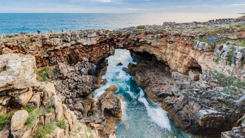A dramatic wide-angle shot of Boca do Inferno in Cascais, Portugal, showing crashing Atlantic waves splashing against the natural limestone rock arch under a cloudy sky.