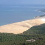 A breathtaking panoramic view of the towering Dune of Pilat in France, showing its sweeping quartz sand slope meeting the Atlantic ocean and green pine forest.