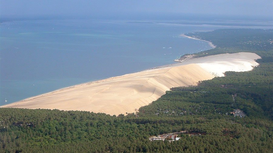 A breathtaking panoramic view of the towering Dune of Pilat in France, showing its sweeping quartz sand slope meeting the Atlantic ocean and green pine forest.