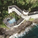 Overgrown cream-colored concrete ruins of an abandoned resort perched high on a tropical limestone cliff overlooking the blue ocean.