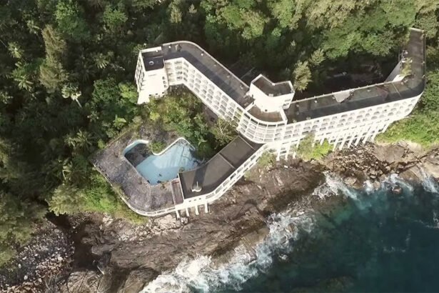 Overgrown cream-colored concrete ruins of an abandoned resort perched high on a tropical limestone cliff overlooking the blue ocean.