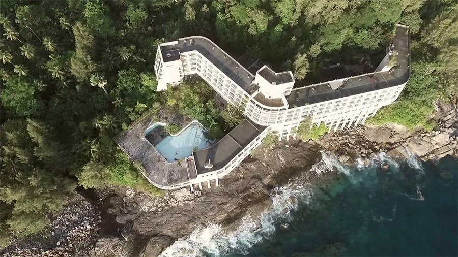 Overgrown cream-colored concrete ruins of an abandoned resort perched high on a tropical limestone cliff overlooking the blue ocean.